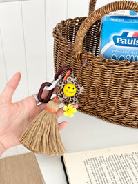 Leopard print flower keychain with a yellow smiley face center, small yellow flower charm, and a tan tassel hanging from chunky dark brown and white links. The keychain is being held in front of a wicker basket containing a milk carton