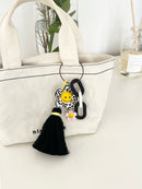 Canvas tote bag with a black-and-white animal print flower keychain attached. The keychain has a yellow smiley face center, a pink mini flower charm, chunky black-and-white links, and a black tassel hanging down, styled as a bag accessory.