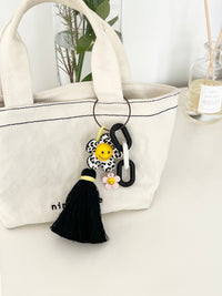 Canvas tote bag with a black-and-white animal print flower keychain attached. The keychain has a yellow smiley face center, a pink mini flower charm, chunky black-and-white links, and a black tassel hanging down, styled as a bag accessory.