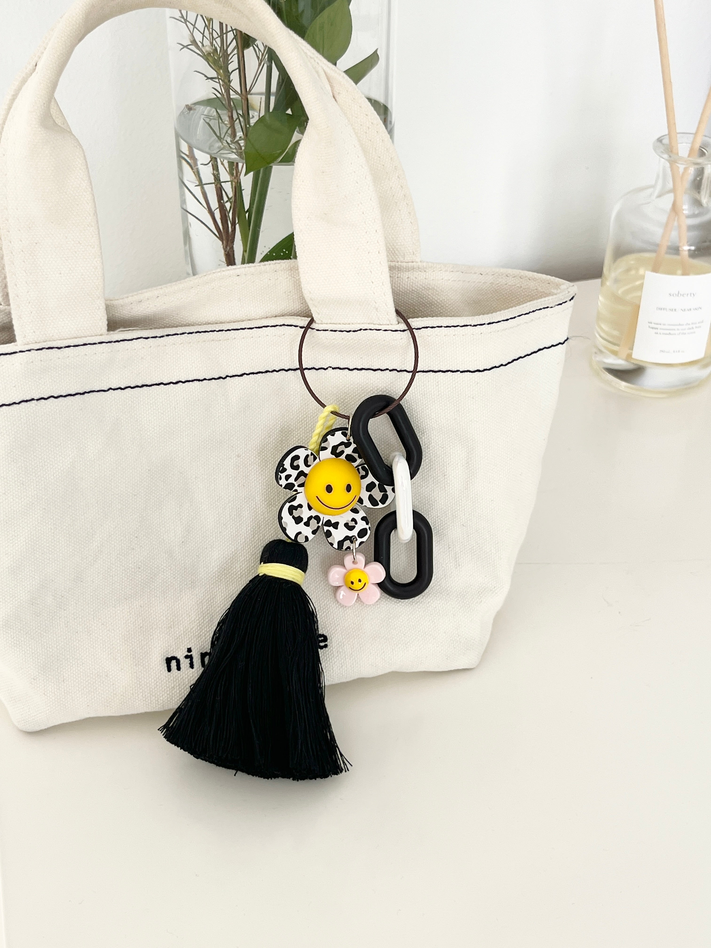 Canvas tote bag with a black-and-white animal print flower keychain attached. The keychain has a yellow smiley face center, a pink mini flower charm, chunky black-and-white links, and a black tassel hanging down, styled as a bag accessory.