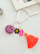 Colorful smiley face keychains with pink rhinestone-covered charms, playful flower accents, and  neon orange tassels, displayed on white surface attached to a bag.