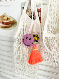 Colorful smiley face keychains with pink rhinestone-covered charms, playful flower accents, and  neon orange tassels, displayed on white surface hanging from a mesh bag.