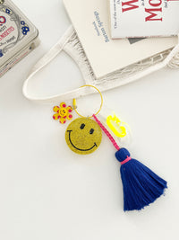Colorful smiley face keychains with yellow rhinestone-covered charms, playful flower accents, and  neon blue tassels, displayed on white surface with hanging from a mesh bag.