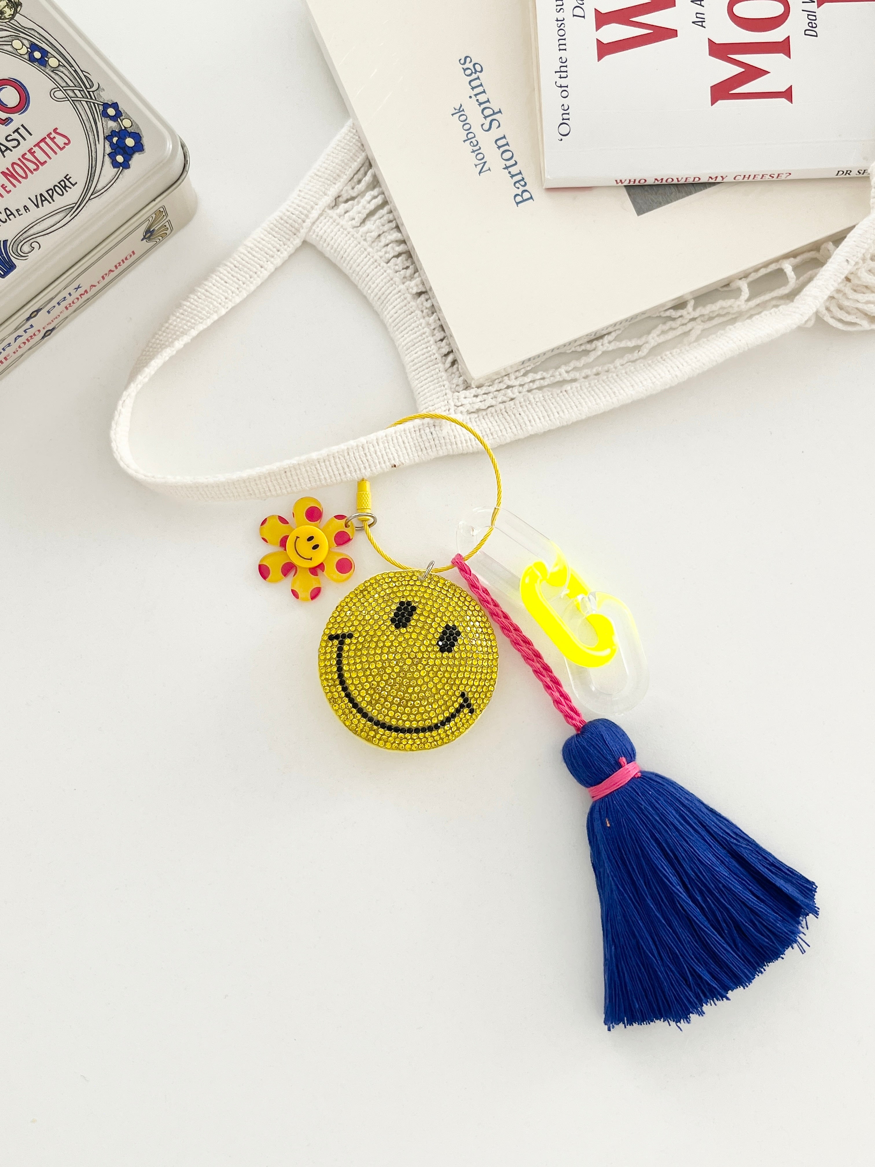Colorful smiley face keychains with yellow rhinestone-covered charms, playful flower accents, and  neon blue tassels, displayed on white surface with hanging from a mesh bag.