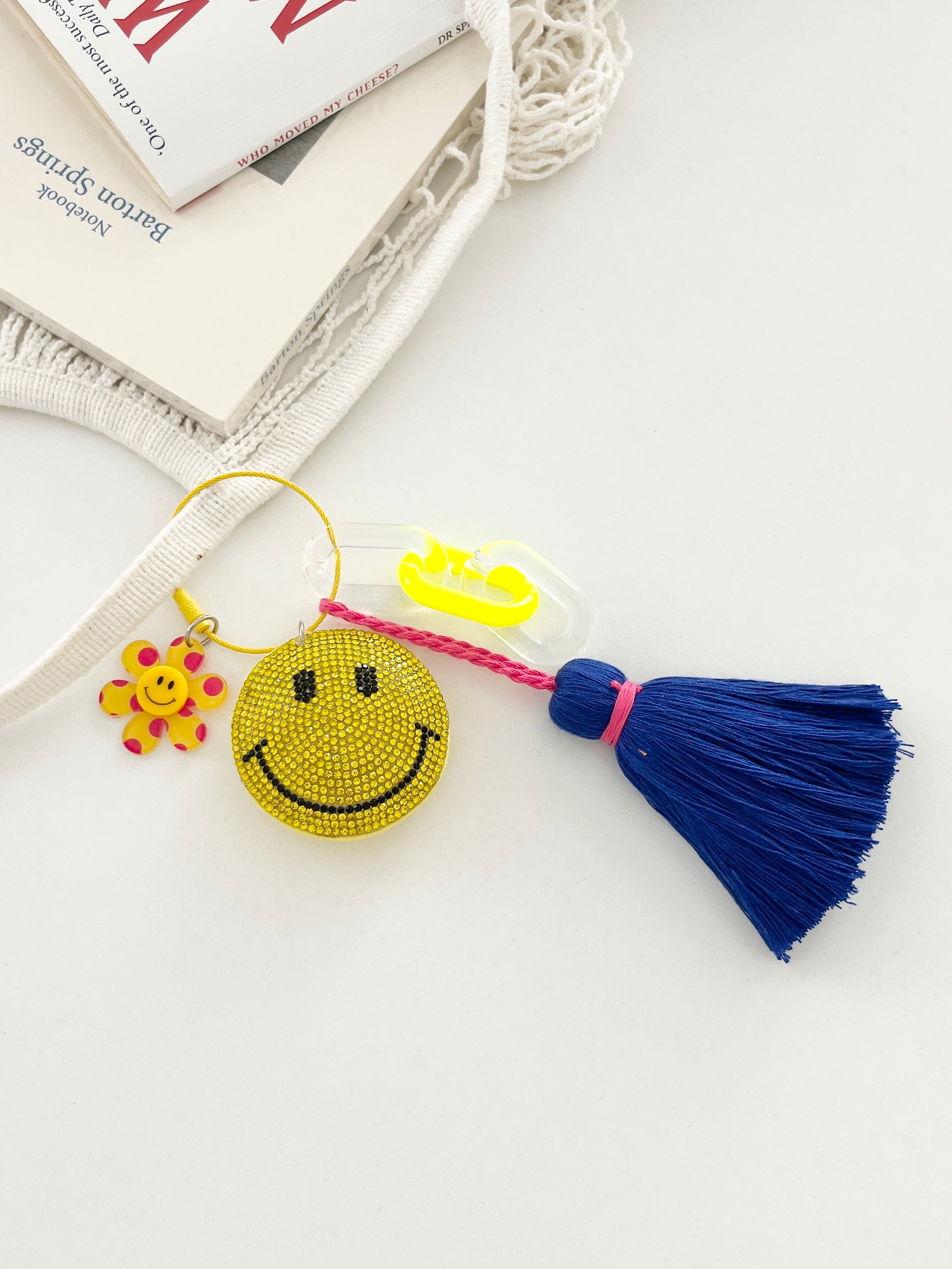 Colorful smiley face keychains with yellow rhinestone-covered charms, playful flower accents, and  neon blue tassels, displayed on white surface with hanging from a mesh bag.