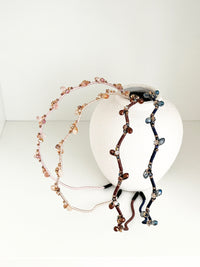 Four crystal hairbands in pale pink, beige, brown, and navy, each with branch-like designs and small crystals, displayed resting on a white round vase.