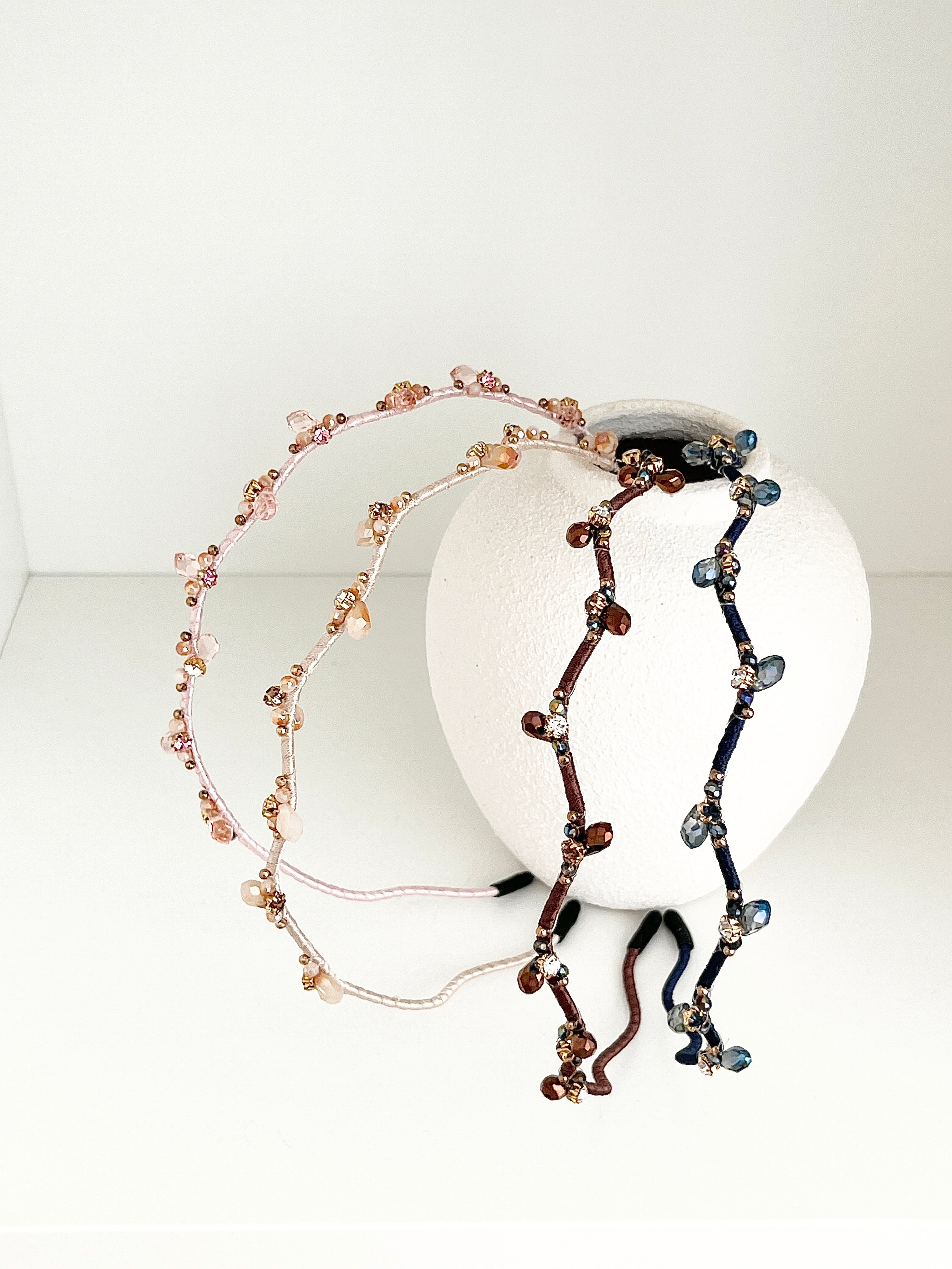 Four crystal hairbands in pale pink, beige, brown, and navy, each with branch-like designs and small crystals, displayed resting on a white round vase.