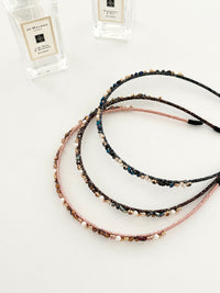 Three slim headbands with crystals, faux pearls, and beads. Includes pink-gold, brown-gold, and dark blue-black designs displayed in a white background with decorative perfume bottles. Lightweight with smooth ends for comfort.