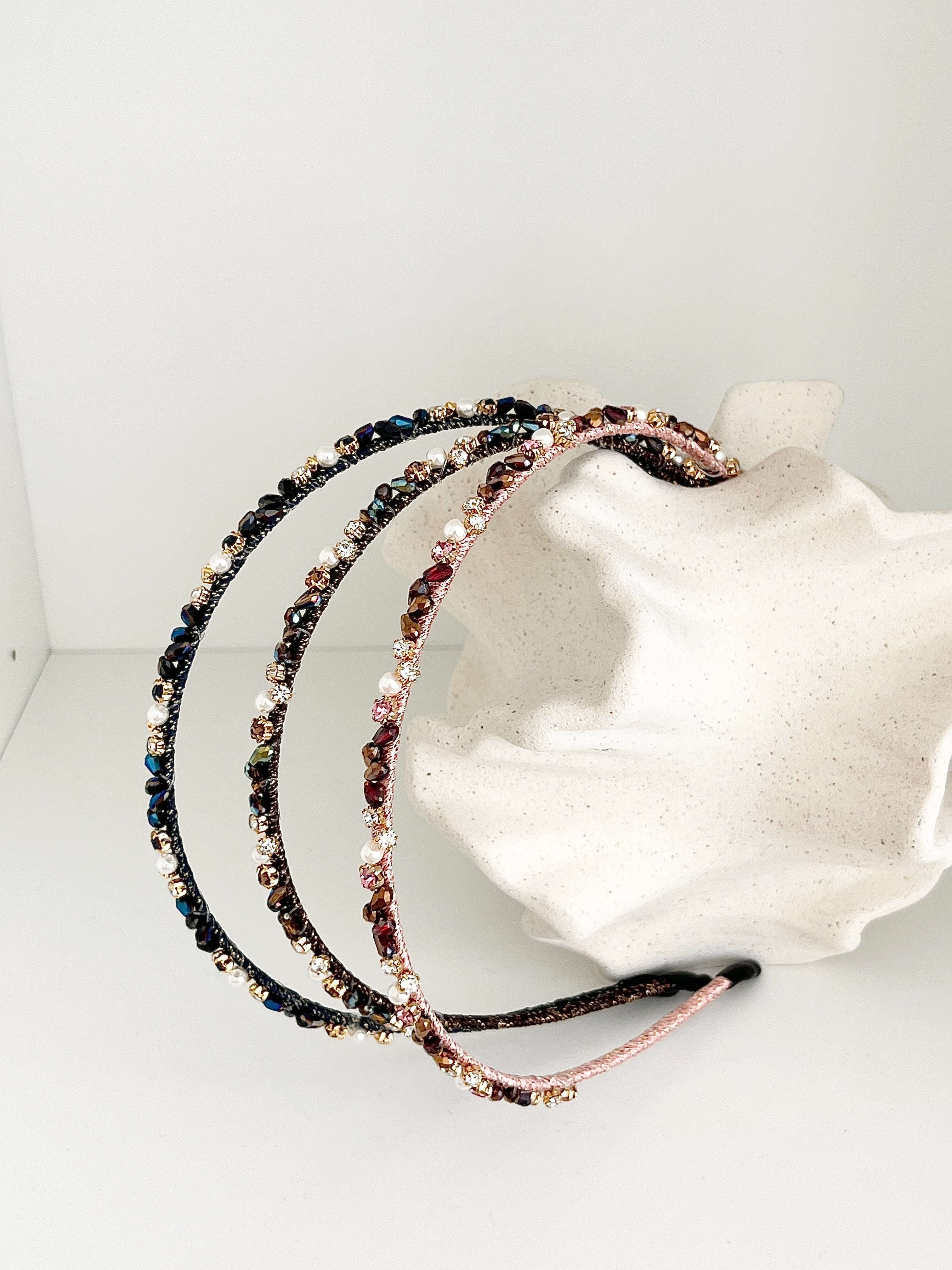 Three slim headbands with crystals, faux pearls, and beads. Includes pink-gold, brown-gold, and dark blue-black designs displayed with decorative stone vase in a white background. Lightweight with smooth ends for comfort.