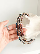 Three slim headbands with crystals, faux pearls, and beads. Includes pink-gold, brown-gold, and dark blue-black designs holding by a hand in a white background. Lightweight with smooth ends for comfort.