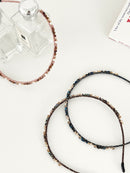 Three slim headbands with crystals, faux pearls, and beads in pink-gold, brown-gold, and dark blue-black. One headband is placed on a flat surface with two clear perfume bottles, while two are displayed in a white surface.