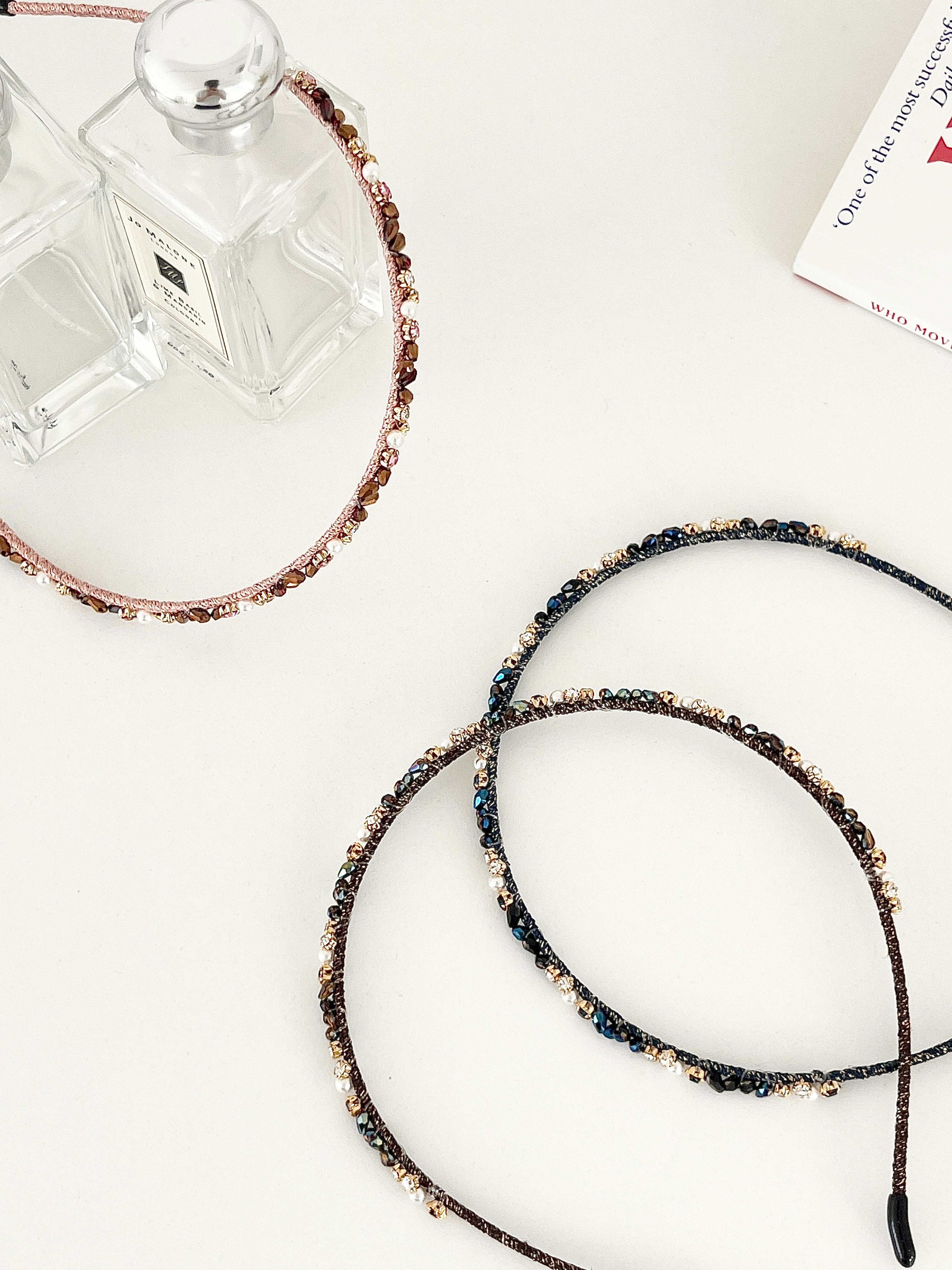 Three slim headbands with crystals, faux pearls, and beads in pink-gold, brown-gold, and dark blue-black. One headband is placed on a flat surface with two clear perfume bottles, while two are displayed in a white surface.