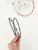 Three slim headbands with crystals, faux pearls, and beads in pink-gold, brown-gold, and dark blue-black. One headband is placed on a flat surface near two clear perfume bottles, while two are held in a hand.