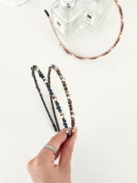 Three slim headbands with crystals, faux pearls, and beads in pink-gold, brown-gold, and dark blue-black. One headband is placed on a flat surface near two clear perfume bottles, while two are held in a hand.