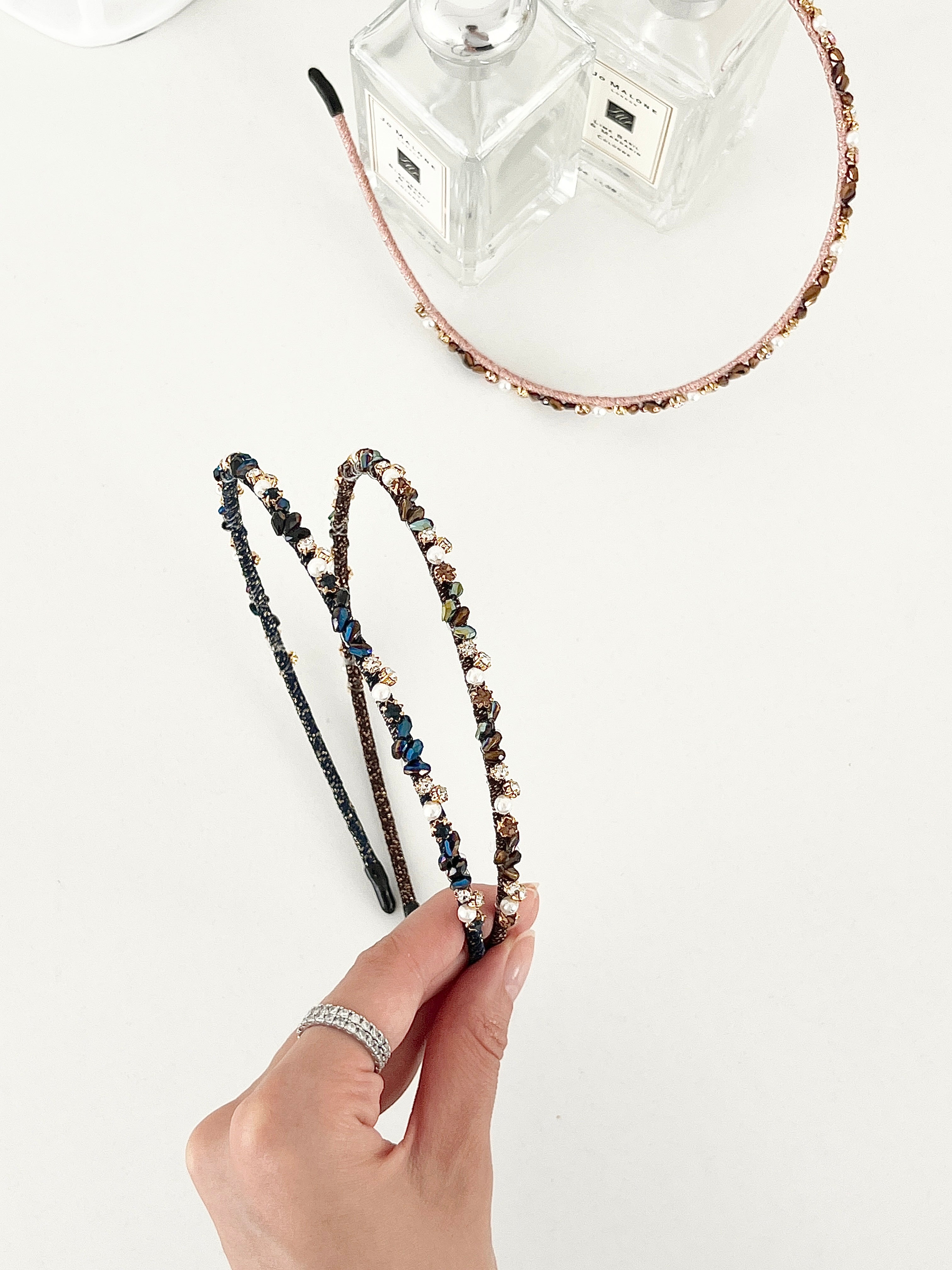 Three slim headbands with crystals, faux pearls, and beads in pink-gold, brown-gold, and dark blue-black. One headband is placed on a flat surface near two clear perfume bottles, while two are held in a hand.