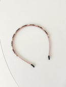 A slim pink headband decorated with small crystals and beads in gold and deep red tones. The band has smooth black tips at each end for comfort. It is placed on a white surface.