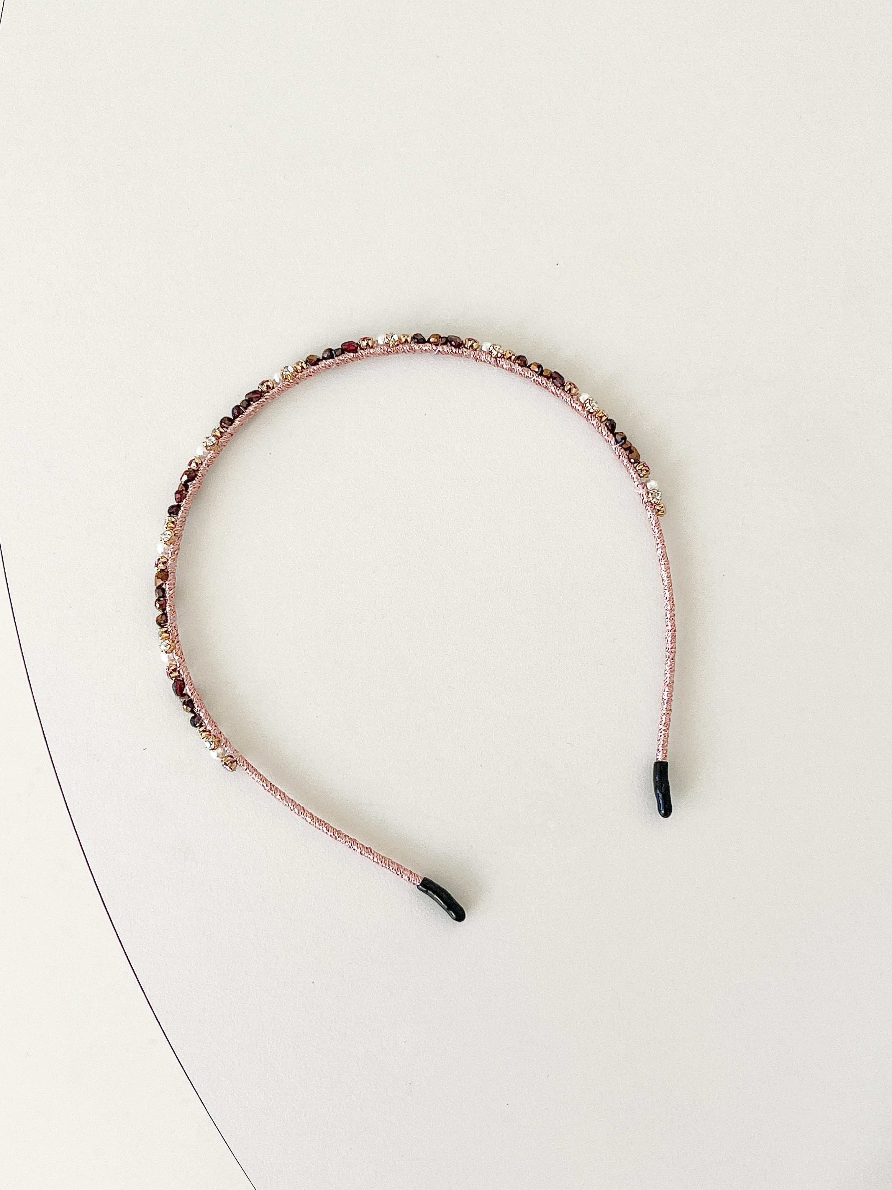 A slim pink headband decorated with small crystals and beads in gold and deep red tones. The band has smooth black tips at each end for comfort. It is placed on a white surface.