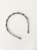 A slim pink headband decorated with small crystals and beads in deep blue, navy and black tones. The band has smooth black tips at each end for comfort. It is placed on a white surface.