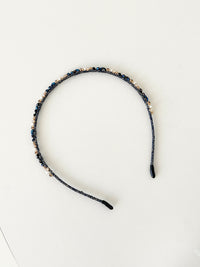 A slim pink headband decorated with small crystals and beads in deep blue, navy and black tones. The band has smooth black tips at each end for comfort. It is placed on a white surface.