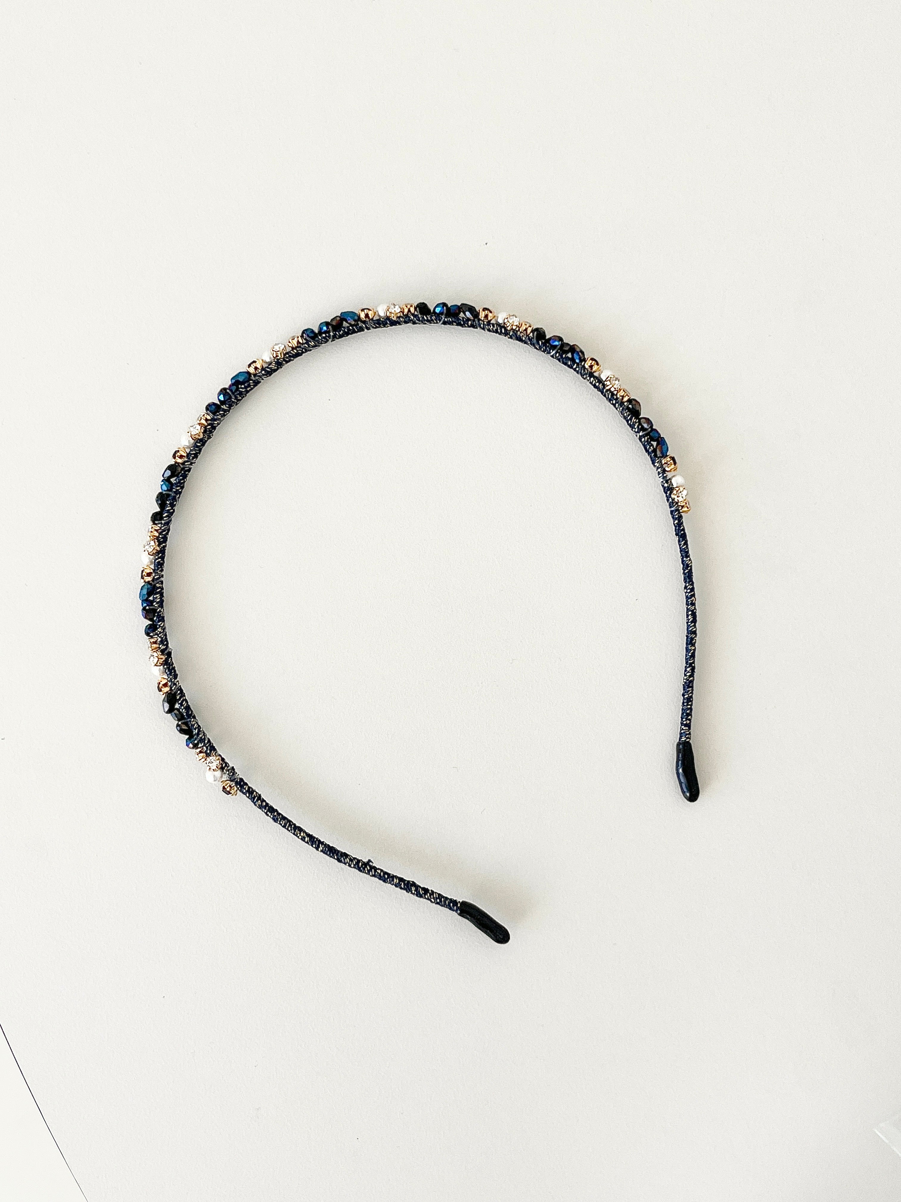 A slim pink headband decorated with small crystals and beads in deep blue, navy and black tones. The band has smooth black tips at each end for comfort. It is placed on a white surface.