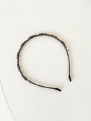 A slim pink headband decorated with small crystals and beads in brown, deep green and white tones. The band has smooth black tips at each end for comfort. It is placed on a white surface.
