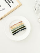 Five slim rectangular hair clips in marbled shades of pink, yellow, jade, dark green, and navy, neatly arranged on a white plate with a framed print and perfume bottles in the background.