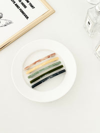 Five slim rectangular hair clips in marbled shades of pink, yellow, jade, dark green, and navy, neatly arranged on a white plate with a framed print and perfume bottles in the background.