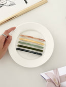 Five slim rectangular hair clips in marbled shades of pink, yellow, jade, dark green, and navy, neatly arranged on a white plate with a framed print with a hand holding the plate.