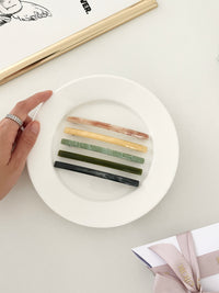 Five slim rectangular hair clips in marbled shades of pink, yellow, jade, dark green, and navy, neatly arranged on a white plate with a framed print with a hand holding the plate.