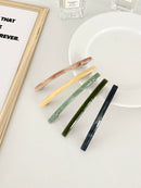 Five slim rectangular hair clips in marbled shades of pink, yellow, jade, dark green, and navy, neatly arranged on a white plate with a framed print and perfume bottles in the background.