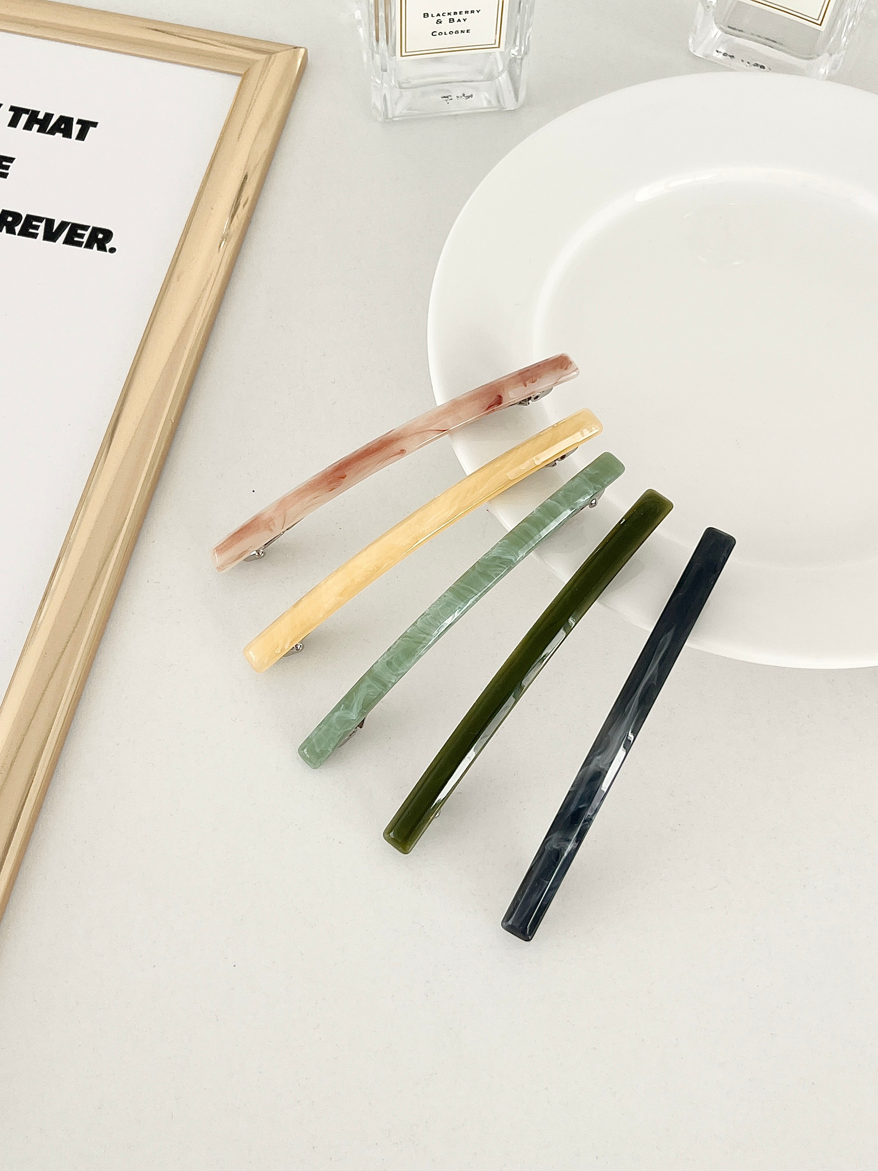 Five slim rectangular hair clips in marbled shades of pink, yellow, jade, dark green, and navy, neatly arranged on a white plate with a framed print and perfume bottles in the background.