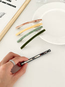 Five slim rectangular hair clips in marbled shades of pink, yellow, jade, dark green, and navy, neatly arranged on a white plate with a framed print with one clip held in hand to display the back clasp.
