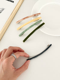 Five slim rectangular hair clips in marbled shades of pink, yellow, jade, dark green, and navy, neatly arranged on a white plate with a framed print with hand holding the navy clip.