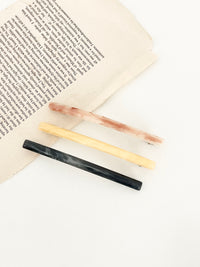 Three slim rectangular hair clips in marbled shades of pink, yellow, and navy, neatly arranged next to an open book on a white plate in white surface.