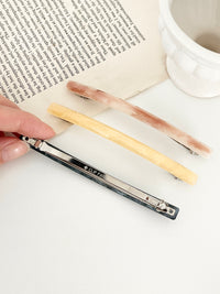 Three slim rectangular hair clips in marbled shades of pink, yellow, and navy, neatly arranged next to an open book on a white plate in white surface with one clip held in hand to display the back clasp.