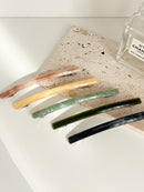 Five slim rectangular hair clips in marbled shades of pink, yellow, jade, dark green, and navy, neatly arranged on a rectangular stone plate with a perfume bottles in the background.