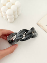 Hand holding a grey hair clip with chain-link design displayed with candle in white background.