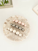 Three pearl-like ball hair clips, in metallic gray, blush pink, and ivory tones, displayed on a stone dish with white surface.