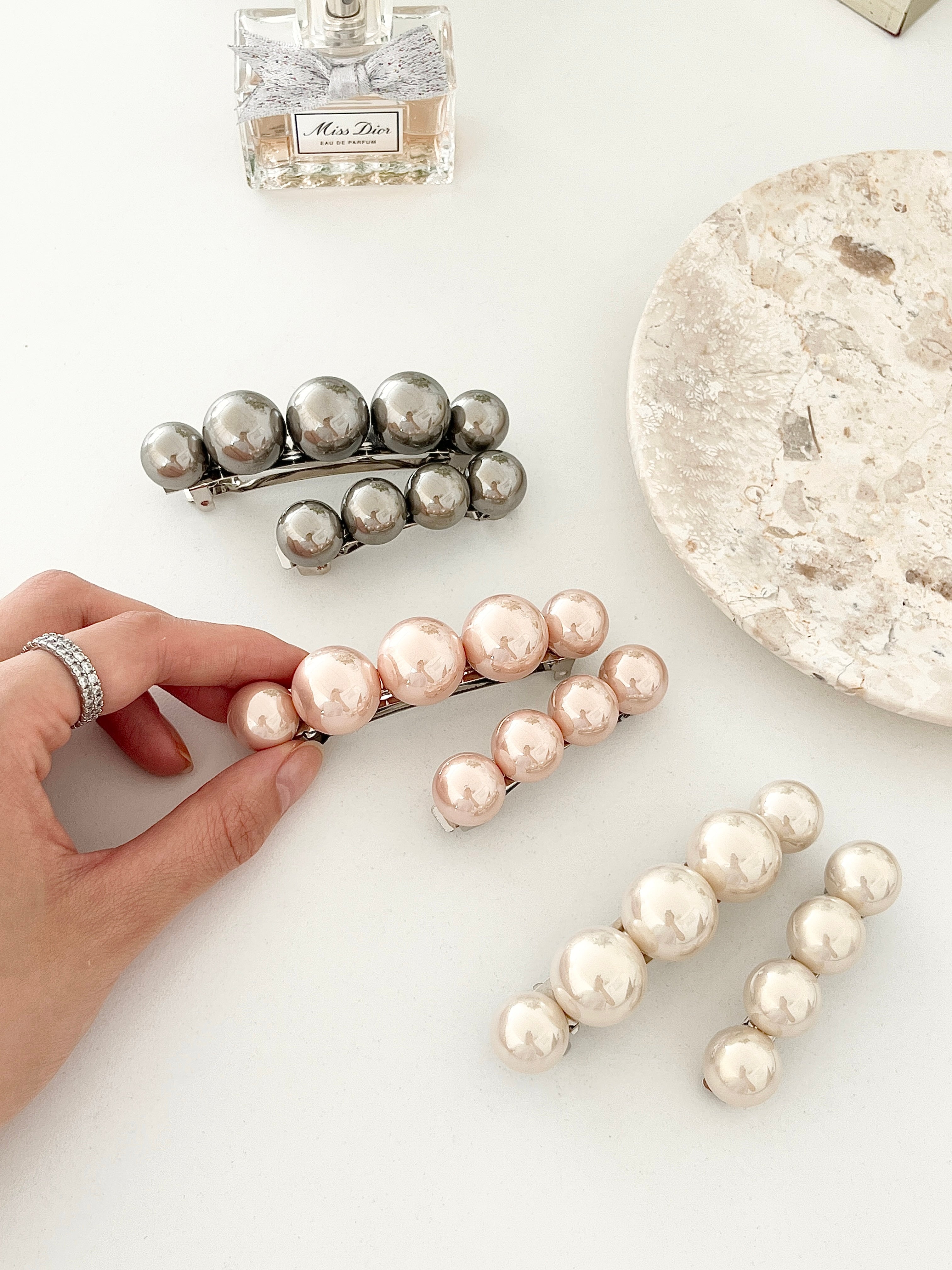 Three matching sets of pearl-like ball hair clips, each set including one large and one small clip, in metallic gray, blush pink, and ivory tones, displayed on a white surface with a hand holding one clip, next to a Miss Dior perfume bottle and a stone dish.