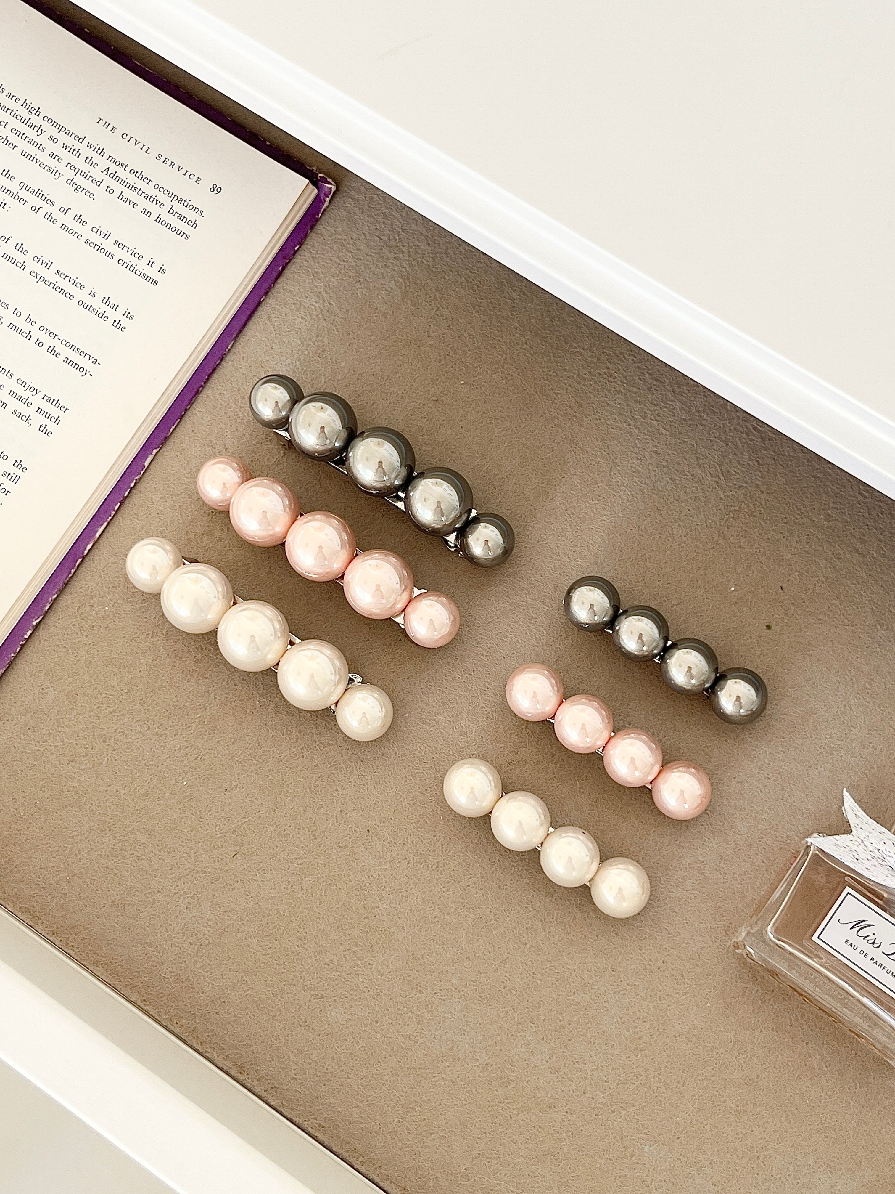 Three sets of pearl-like ball hair clips, each set including one large and one small clip, in metallic gray, blush pink, and ivory tones, neatly arranged in a drawer next to an open book and a Miss Dior perfume bottle.