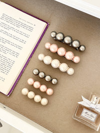 Three sets of pearl-like ball hair clips, each set including one large and one small clip, in metallic gray, blush pink, and ivory tones, neatly arranged in a drawer next to an open book and a Miss Dior perfume bottle.