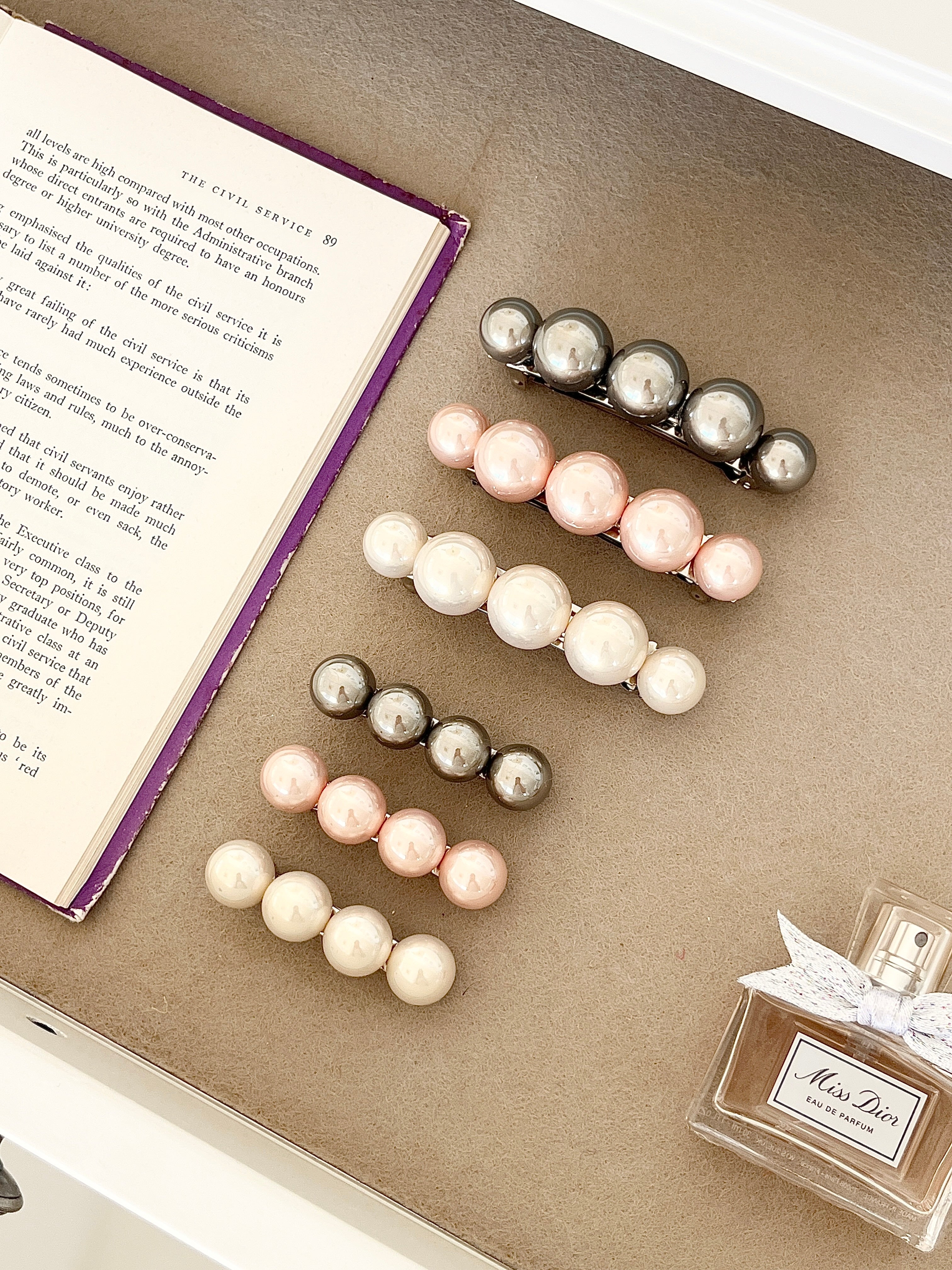 Three sets of pearl-like ball hair clips, each set including one large and one small clip, in metallic gray, blush pink, and ivory tones, neatly arranged in a drawer next to an open book and a Miss Dior perfume bottle.