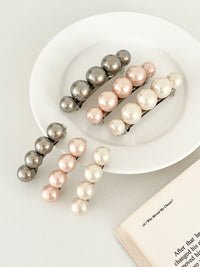 Three sets of pearl-like ball hair clips, each set including one large and one small clip, in metallic gray, blush pink, and ivory tones, neatly arranged on a white plate next to an open book.