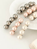Three sets of pearl-like ball hair clips, each set including one large and one small clip, in metallic gray, blush pink, and ivory tones, neatly arranged on a white plate next to an open book.