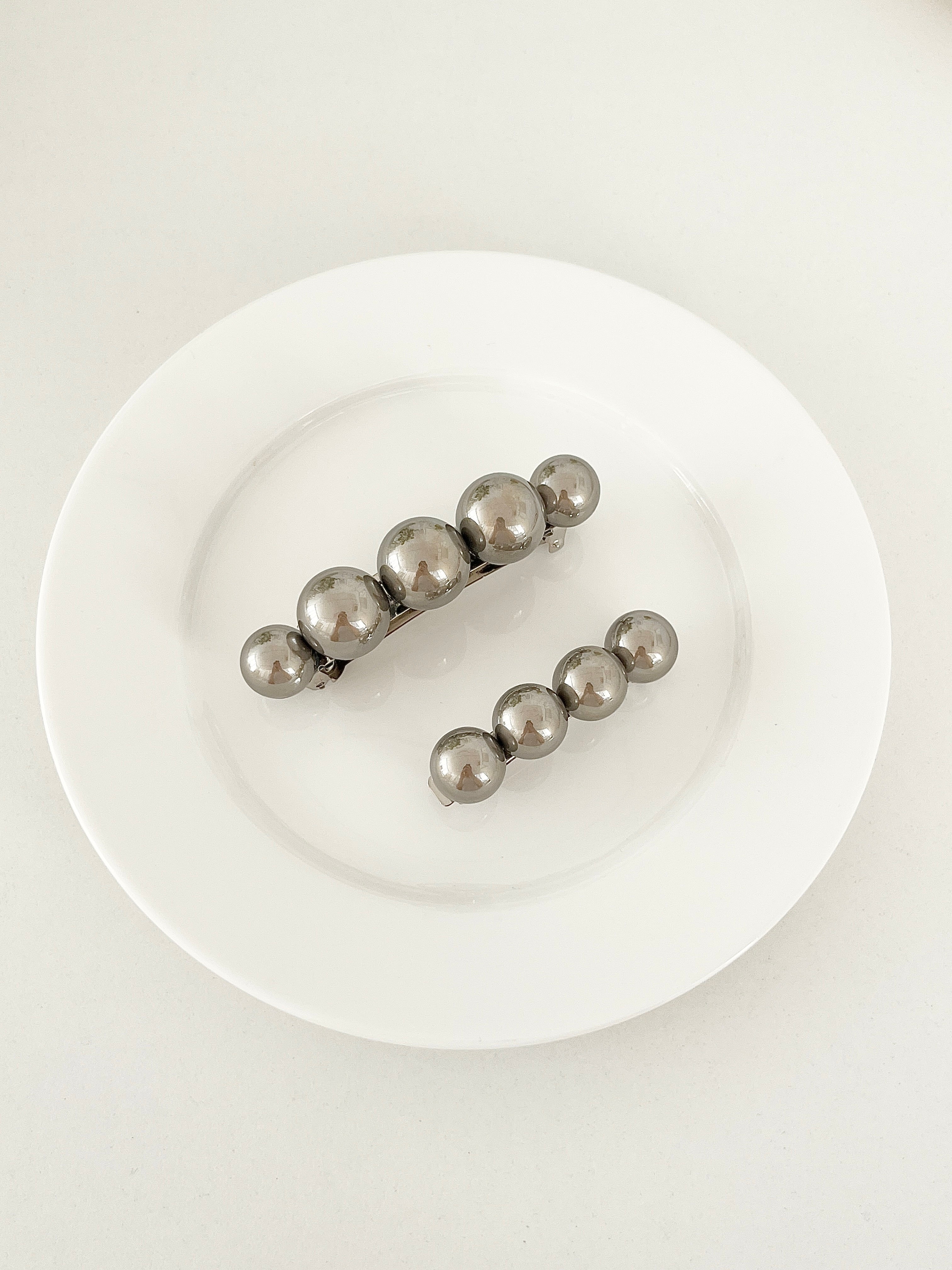 One matching sets of pearl-like ball hair clips, each set including one large and one small clip, in metallic gray, displayed on a white plate with white surface.