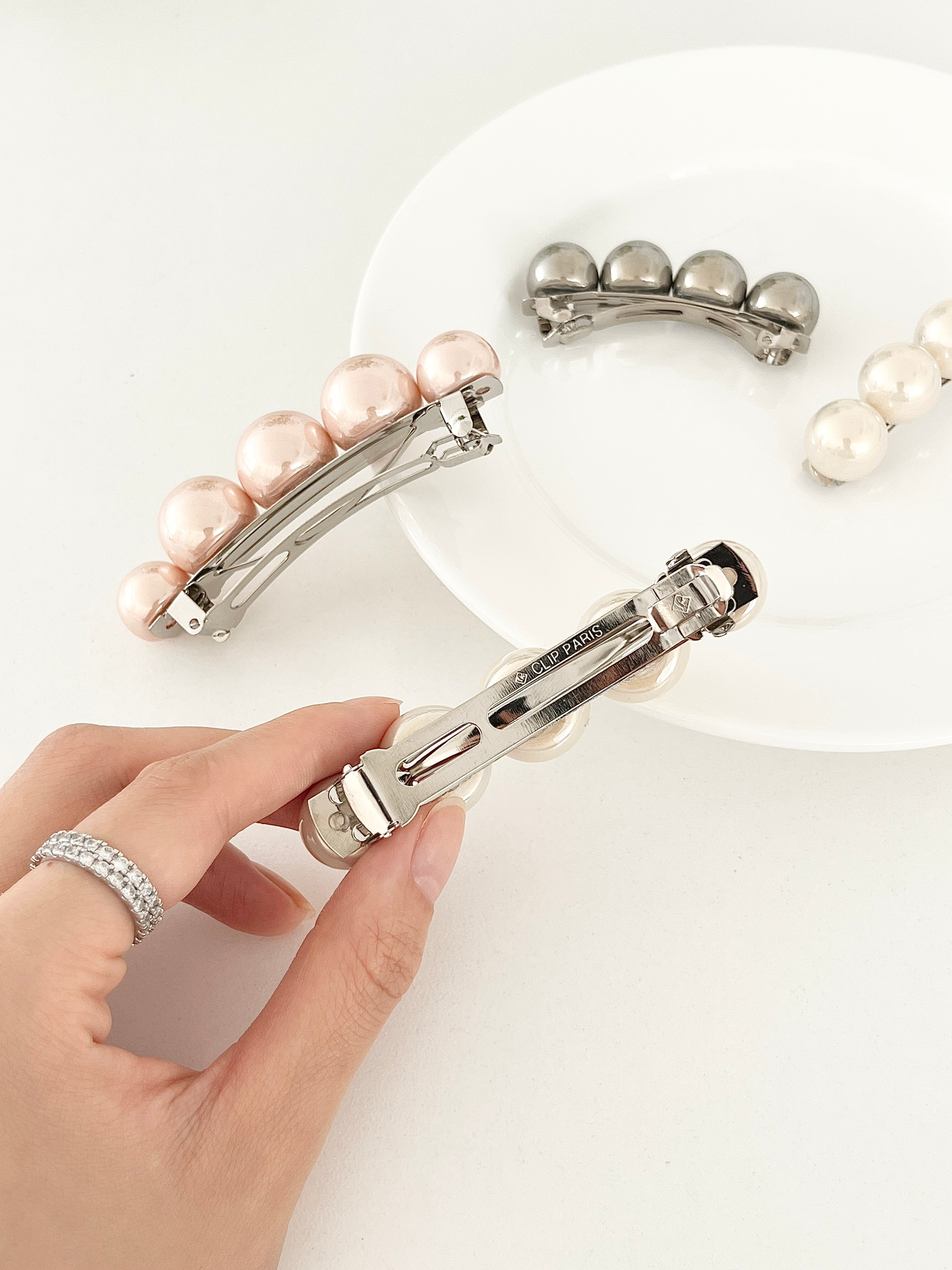 Four pearl-like ball hair clips, displayed on a white plate with white surface with held in hand to display the back clasp.