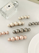 Three sets of pearl-like ball hair clips, each set including one large and one small clip, in metallic gray, blush pink, and ivory tones, neatly arranged on a white plate next to perfume bottle.