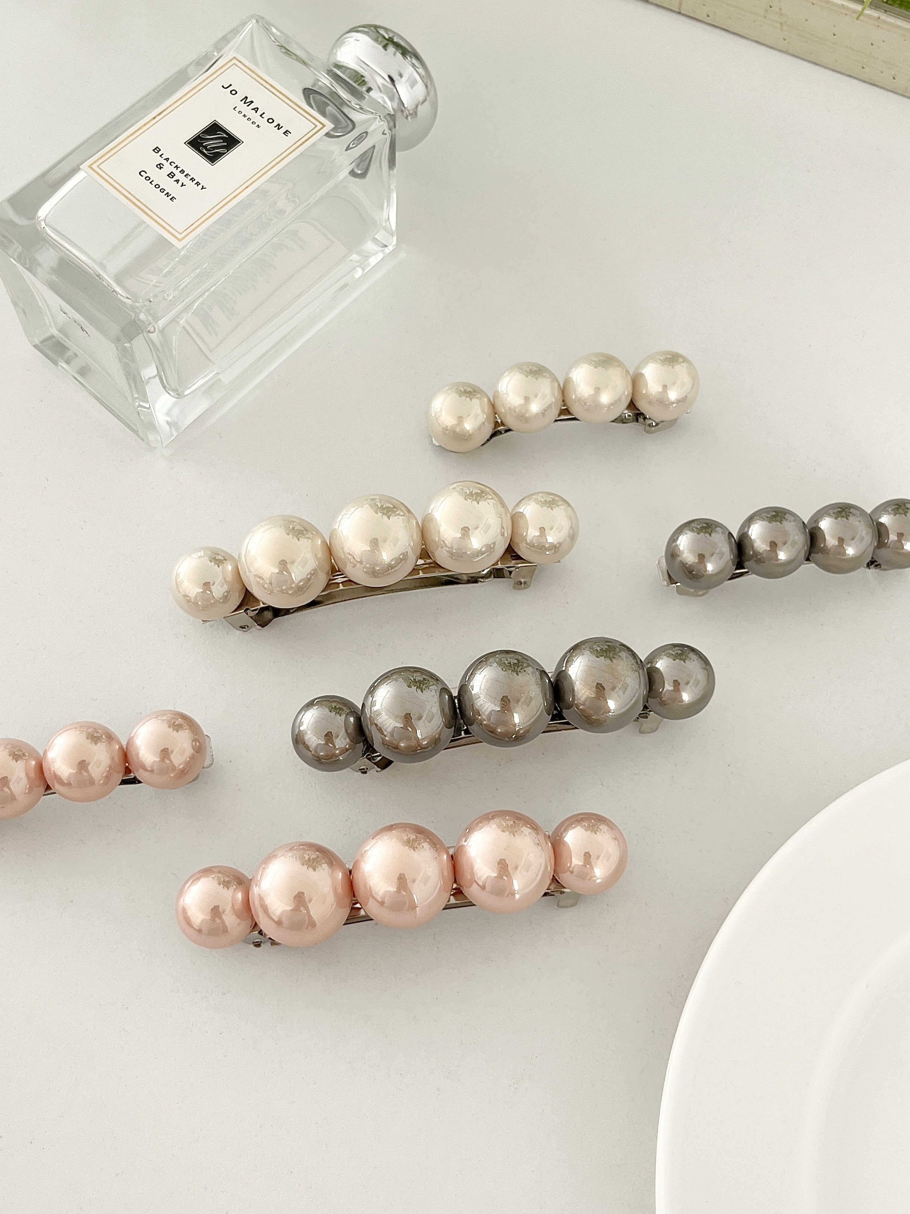 Three sets of pearl-like ball hair clips, each set including one large and one small clip, in metallic gray, blush pink, and ivory tones, neatly arranged on a white plate next to perfume bottle.