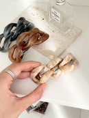 Three chunky chain-link hair clips in gray, brown, and beige tones, displayed on a stone tile next to a Chanel No. 5 perfume bottle. Hand hold a beige chain-link clip on a photo.