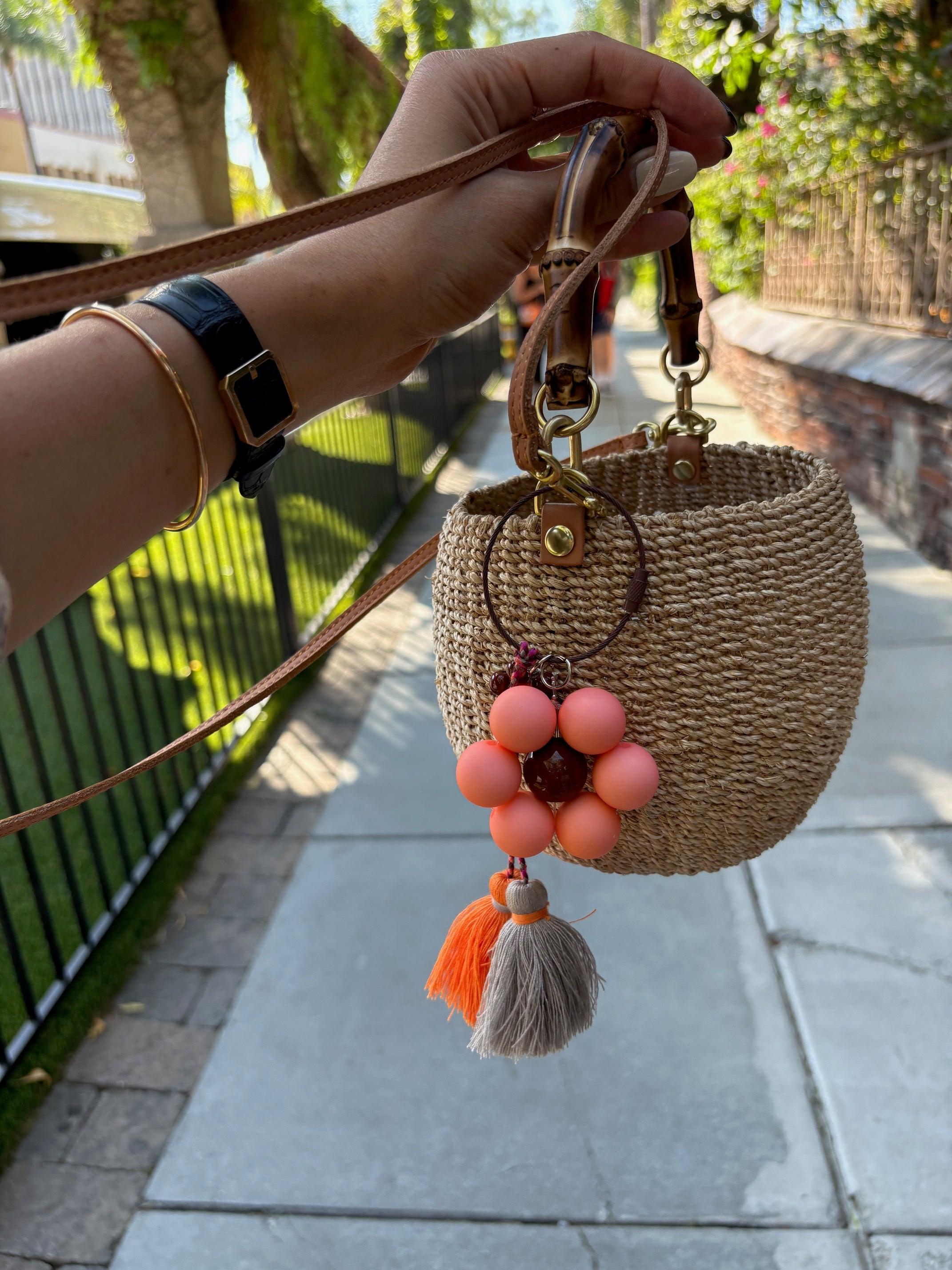 A hand is holding a woven straw handbag with a flower keychain attached. The keychain has a peach flower with a brown center and tassels in gray and orange. 